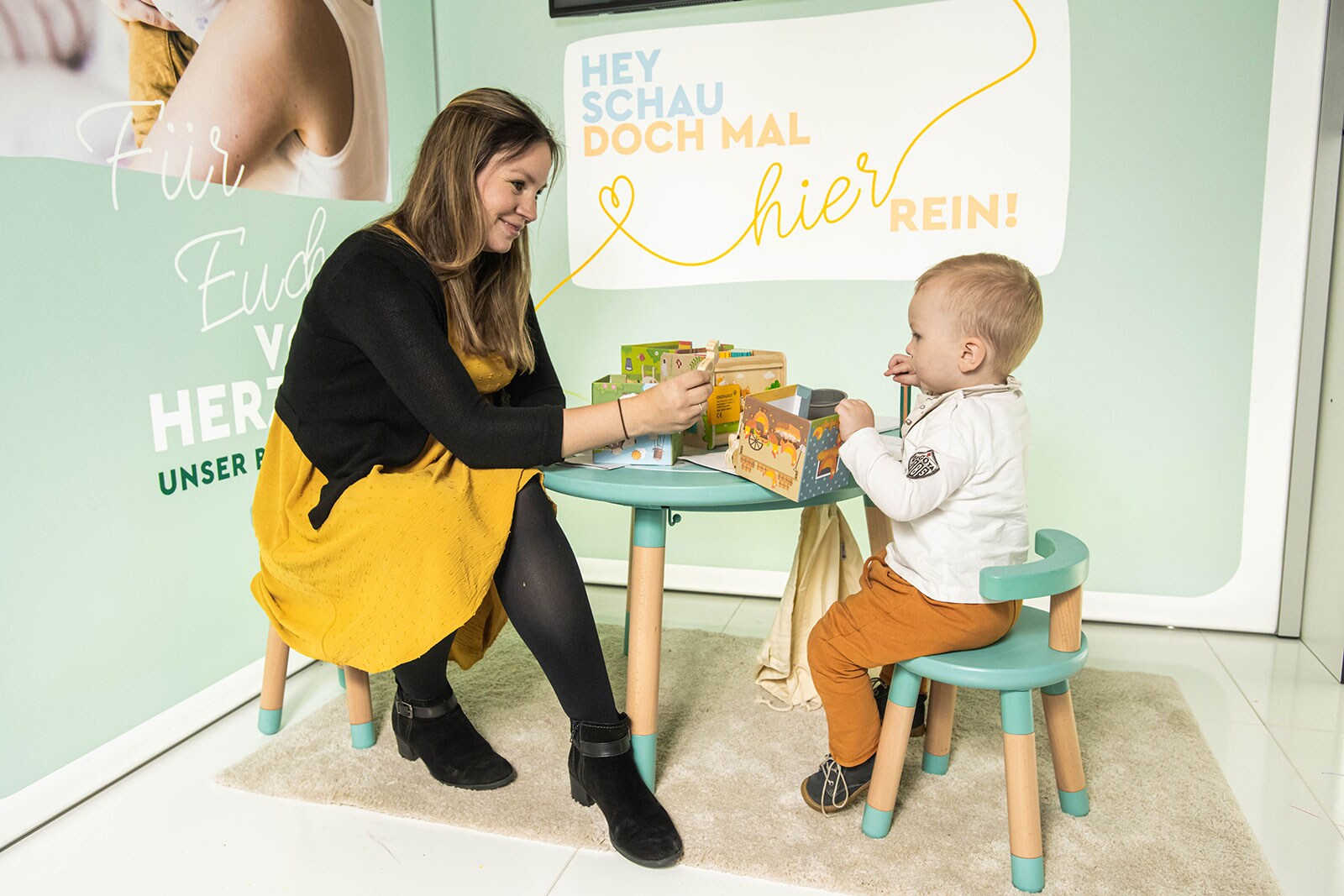 A woman in a yellow dress sits with a toddler at a children&#39;s play table.
