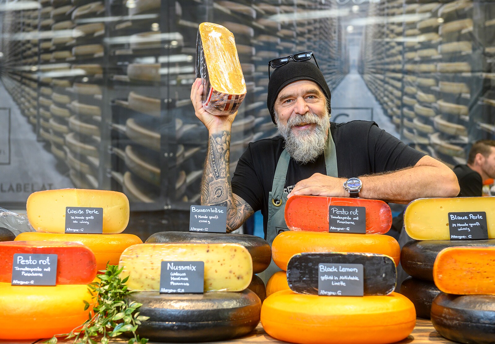 A man holds up half a wheel of cheese. There are many different types of cheese on the counter in front of him