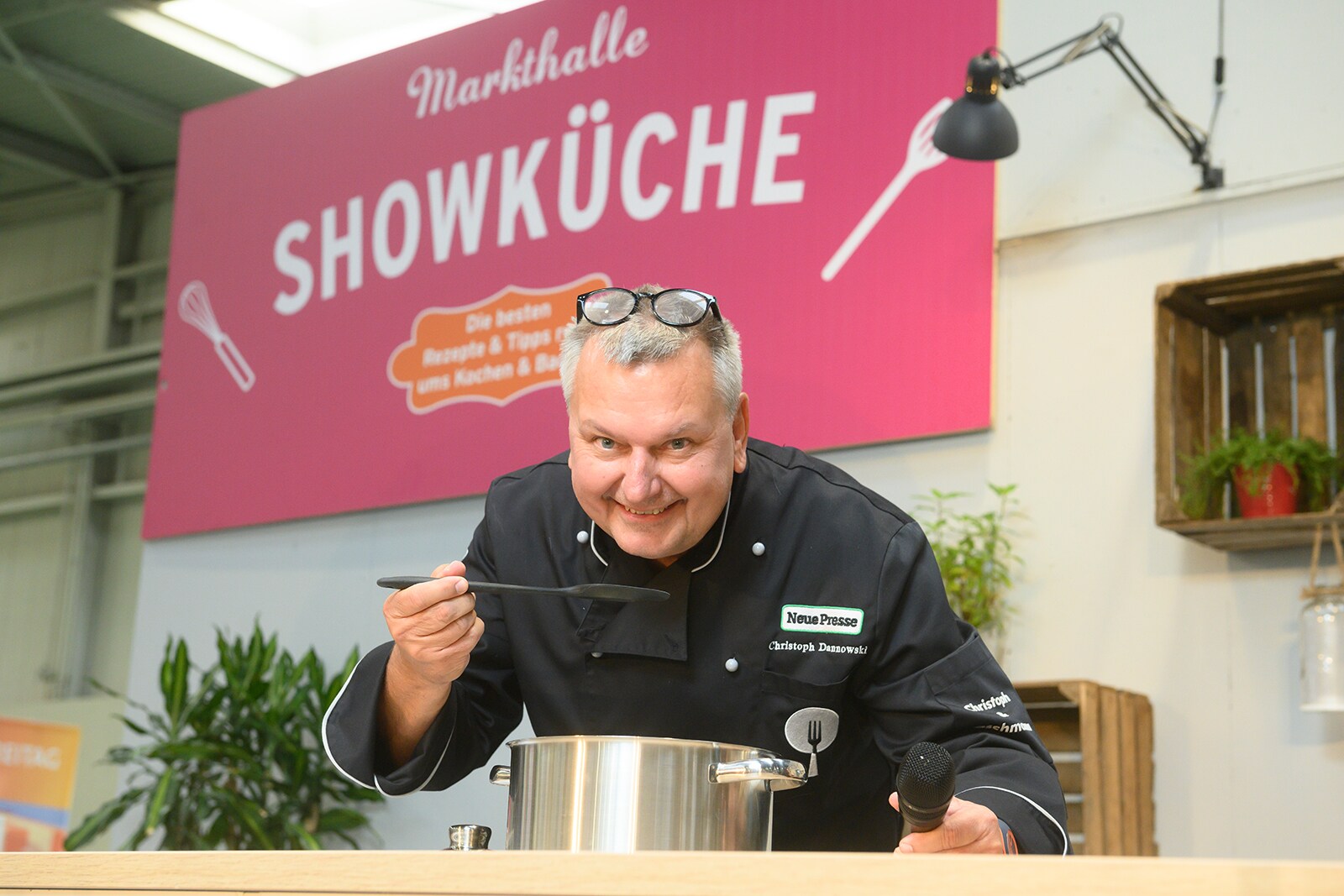 Presenter Christoph Dannowski leans over the counter of the show kitchen and tastes from a pot
