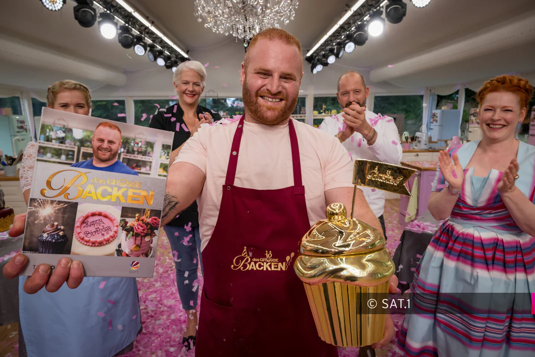 Raheem Haidar is one of the hottest young confectioners in Germany. He stands on the set of the Sat 1 baking show ‘Das grosse Backen’ and holds his prize up to the camera. In the background are his fellow contestants and presenter Enie van de Meiklokjes
