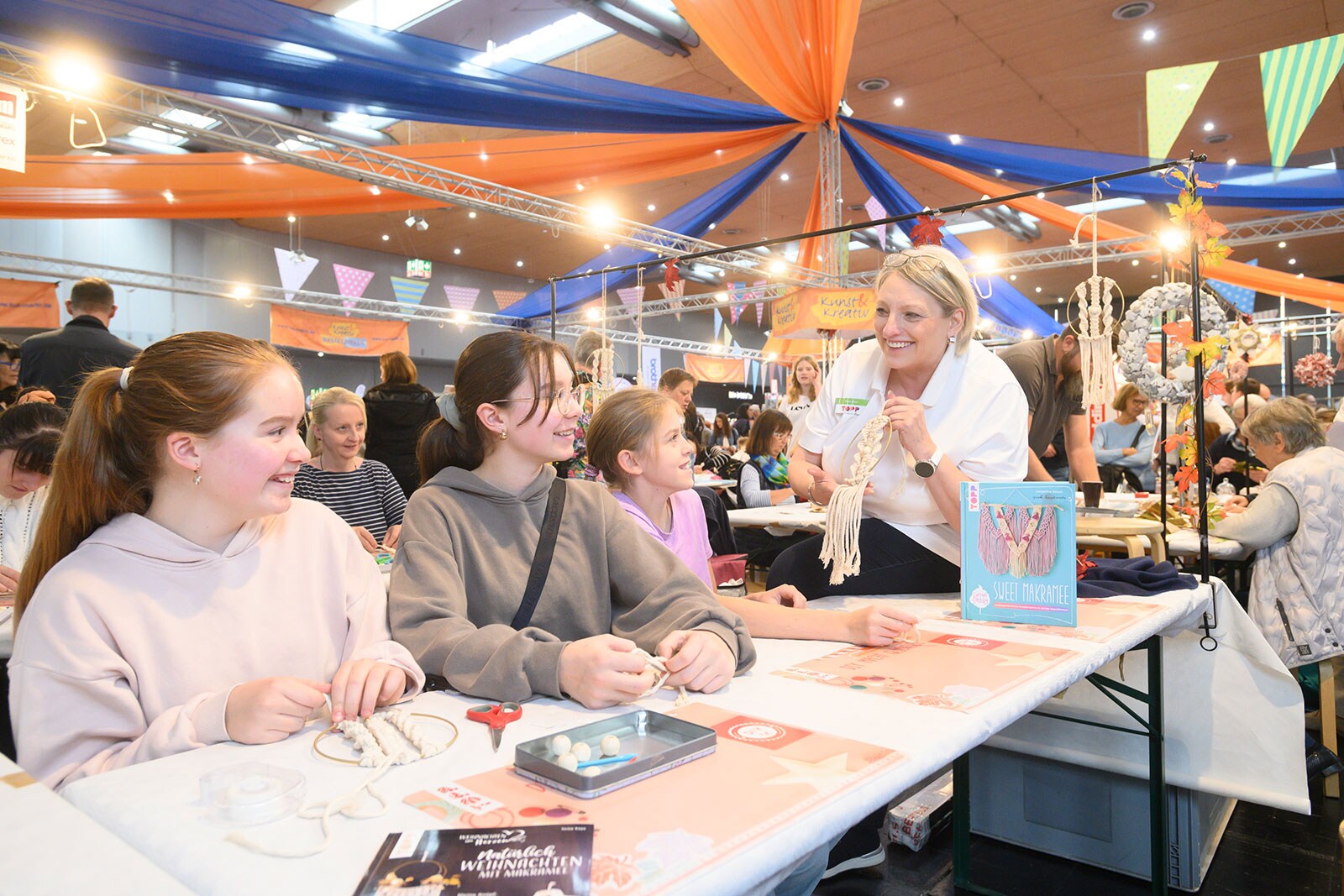 Three young girls take part in a macramé workshop while a woman shows them what the result can look like.