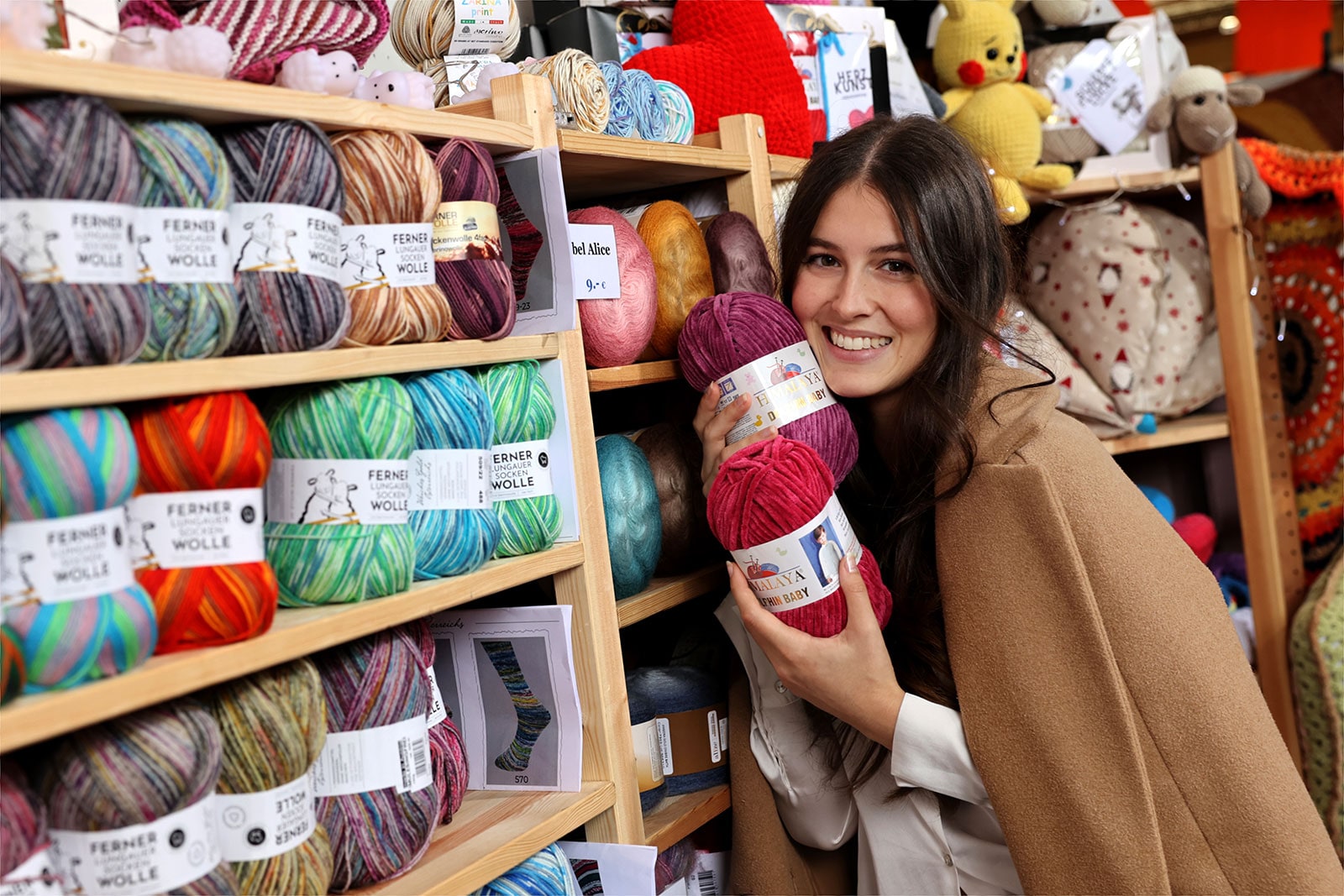 Eine Junge Frau steht vor einem Holzregal mit bunten Wollknäulen. In den Händen hält sie ein rotes und ein beerenfarbenes Knäul