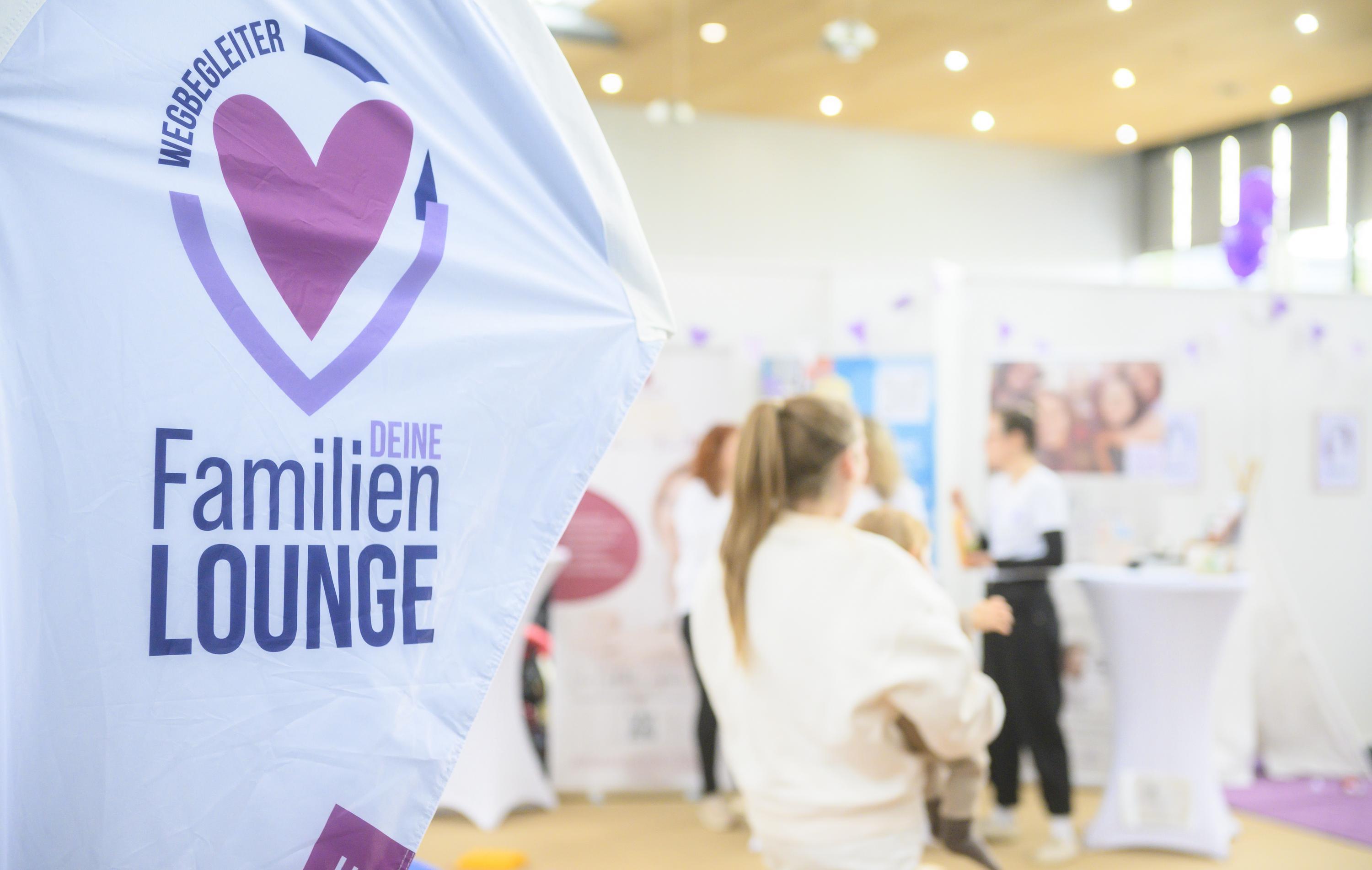 In the background of the infa Messe, a family event, a group of people is gathered at a booth. One person is holding a child while talking to others. In the foreground, a banner with the &#34;Familien Lounge&#34; logo is visible. The booth is bright and friendly, with white tablecloths and purple decorations.